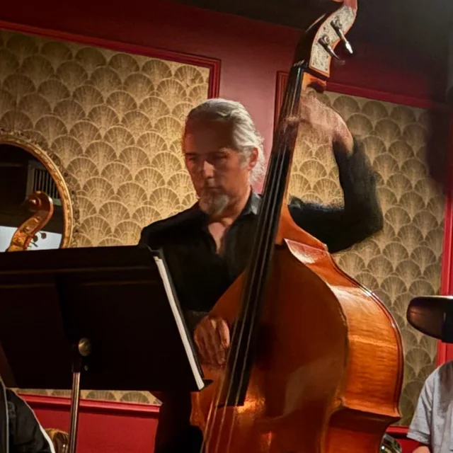 Hoai Nguyen playing his upright bass in a red-walled club, music stand in front of him.