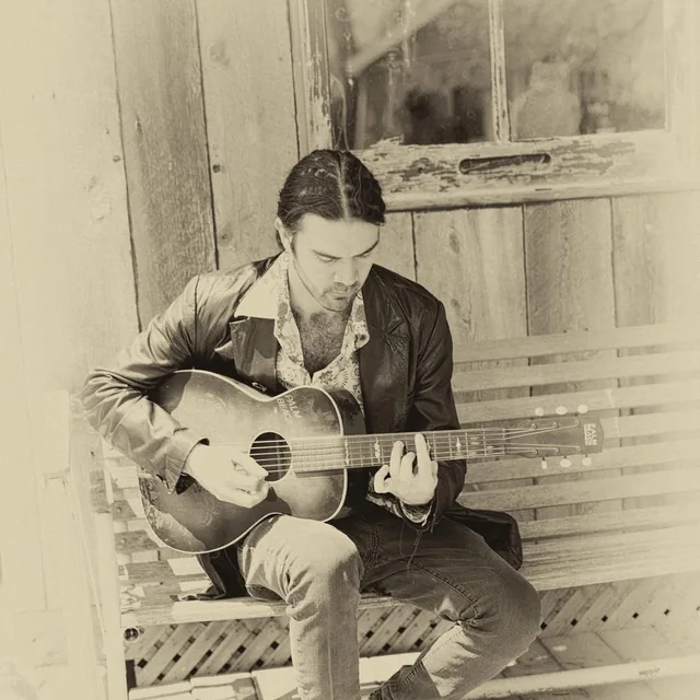 Luke Robertson seated on a porch bench with an acoustic guitar, in a sepia-toned photograph.