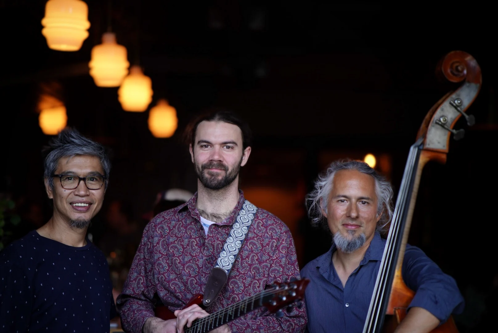 The Railway Trio at night under warm café lights: Gop Intarachot, Luke Robertson holding a red electric guitar, and Hoai Nguyen with his upright bass.